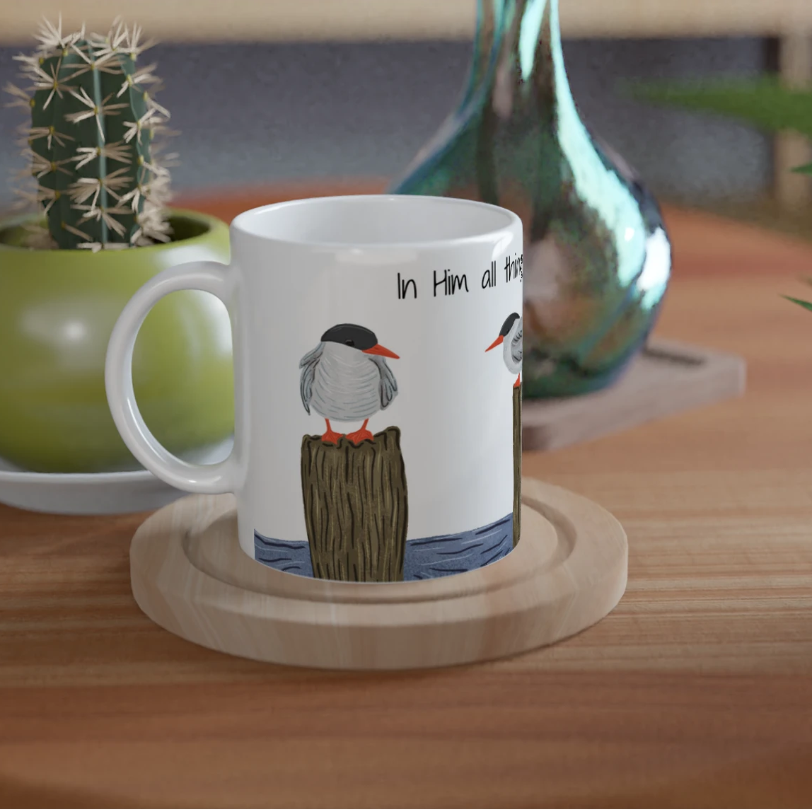 A white ceramic mug with a printed illustration of two birds on a wooden post and the text 'In Hain, all the time' sits on a round wooden coaster on a round wooden table. The scene is styled with a small potted cactus and other home decor in the background.