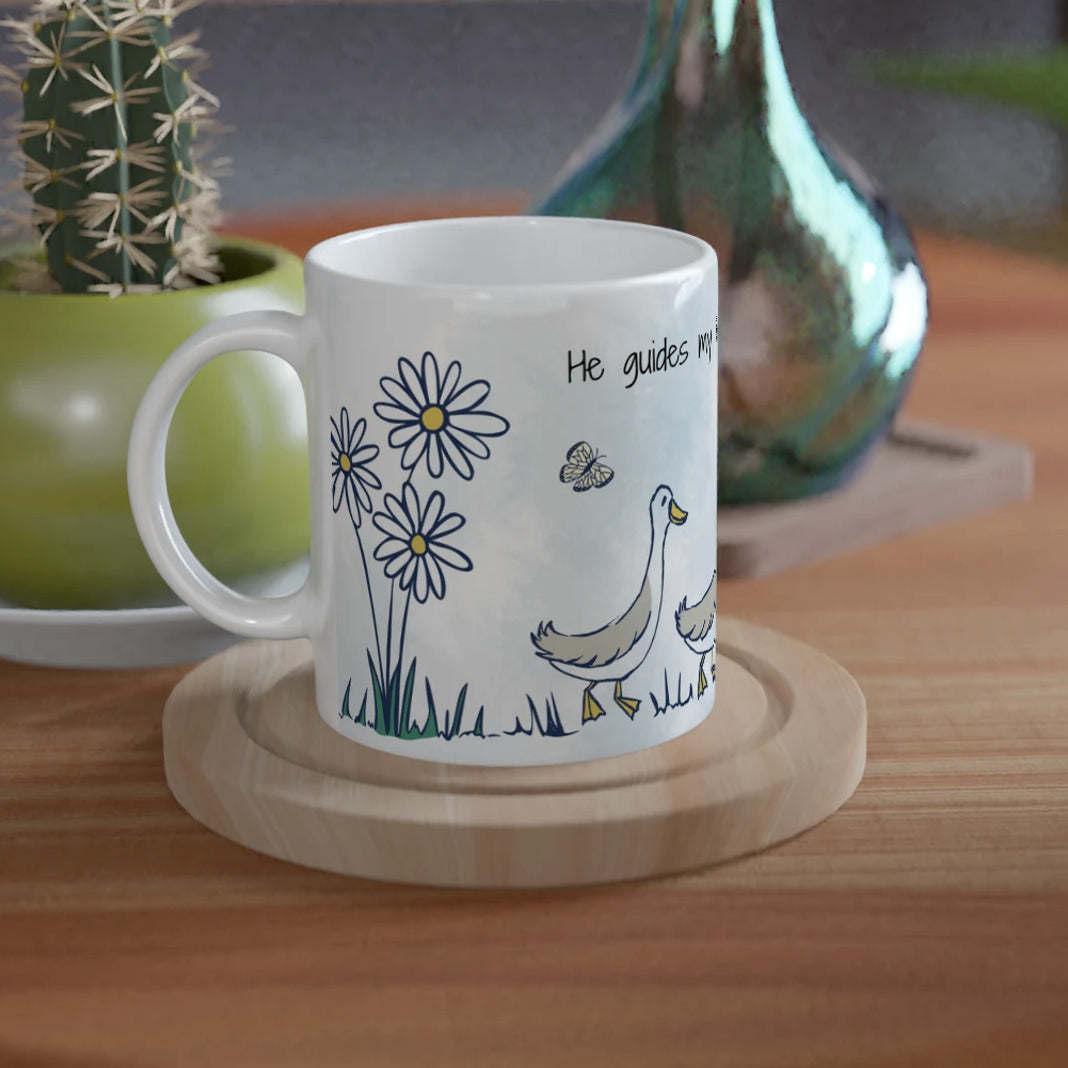 A white ceramic mug with a blue and green duck and flower illustration sits on a round wooden coaster on a wooden table, styled with a small potted cactus and a decorative vase in the background.