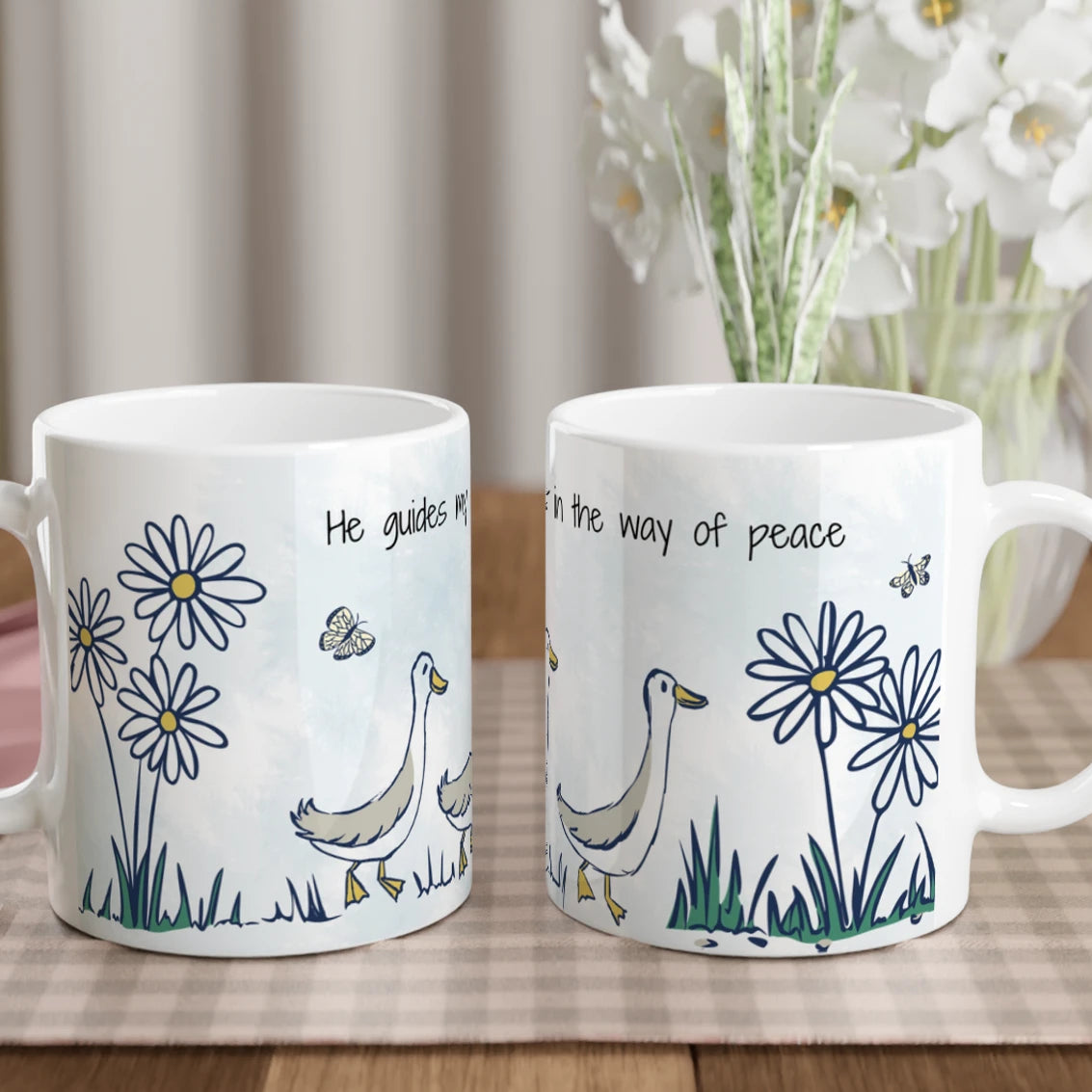 Two white ceramic mugs with a blue and green duck and flower design, displayed side by side on a checkered table mat with a vase of flowers in the background.