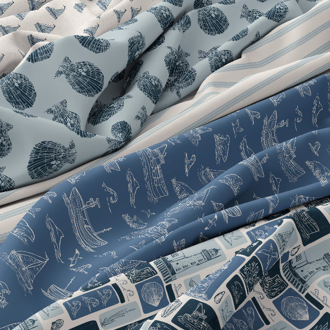 A close-up of draped bedding or fabric with multiple nautical-themed prints: one light blue with dark blue seashell motifs, one medium blue with white ship sketches, and one white with blue stripes and blue ship blocks.