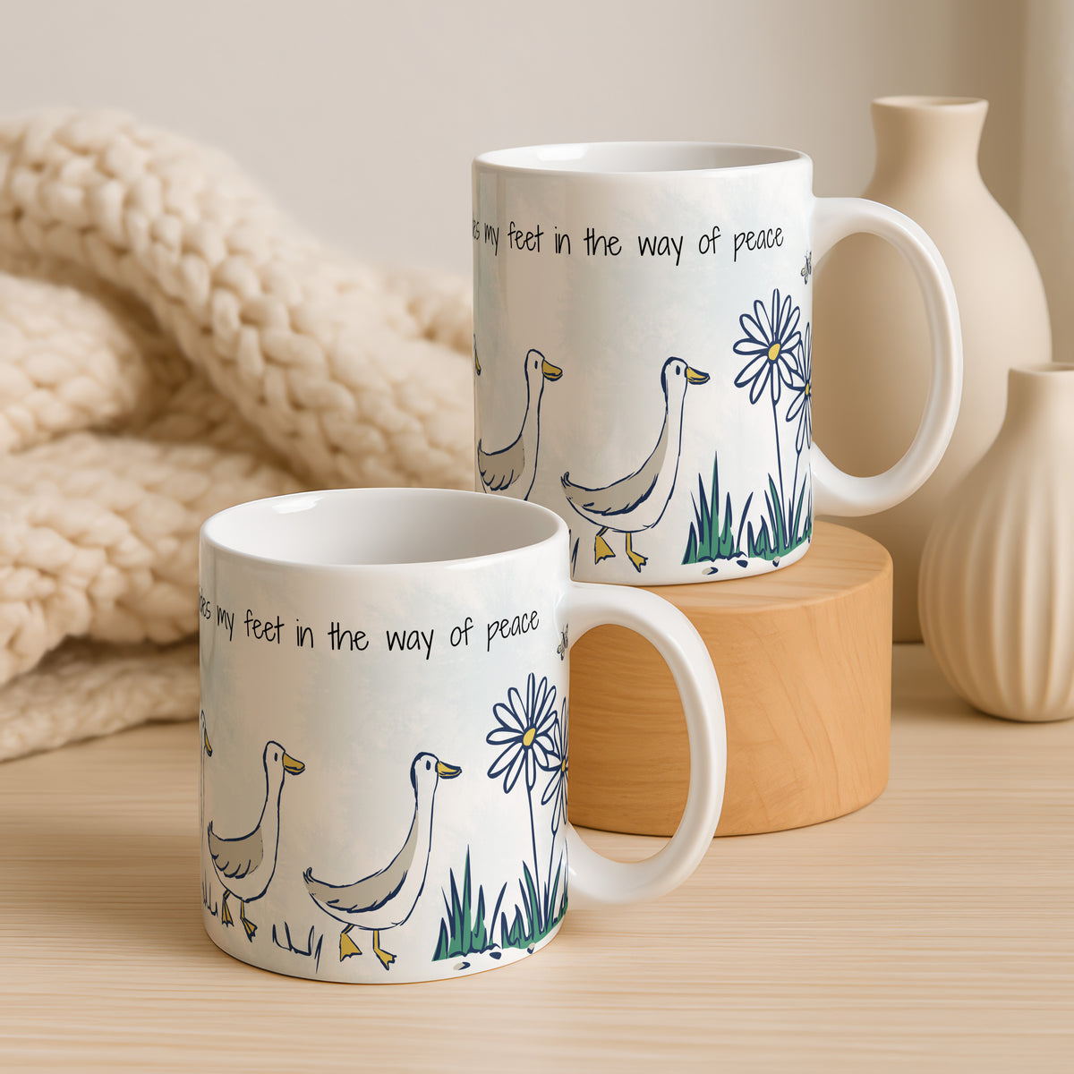 Two white ceramic mugs with a printed design of walking geese, blue flowers, and black text, styled on a wooden surface with a chunky knit blanket and neutral ceramic vases in the background.