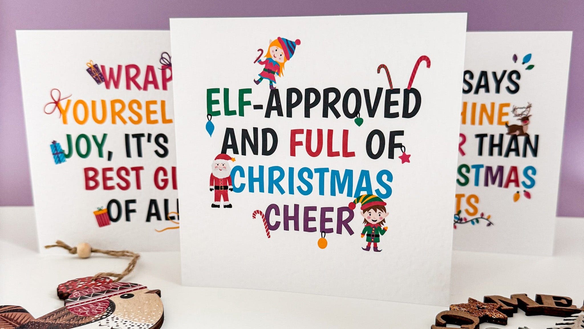 Three Christmas-themed cards with elf illustrations and festive text on a white surface with a purple background.