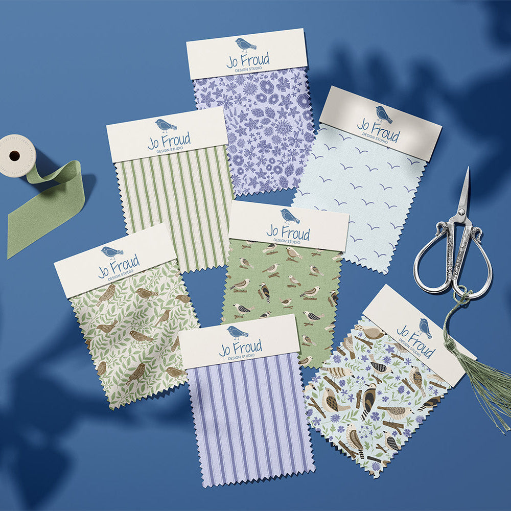 Seven fabric swatch samples from Jo Froud are arranged on a blue background with a spool of ribbon, silver scissors, and a tassel. The swatches display various patterns, including stripes, florals, and birds in shades of purple, green, beige, and blue.