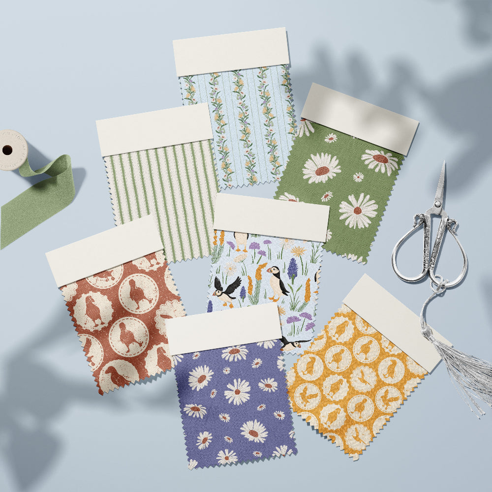 Seven fabric swatch cards with different patterns (stripes, florals, silhouettes) are arranged on a light blue background with a spool of green thread, silver scissors, and a white tassel. The cards have white tabs at the top.