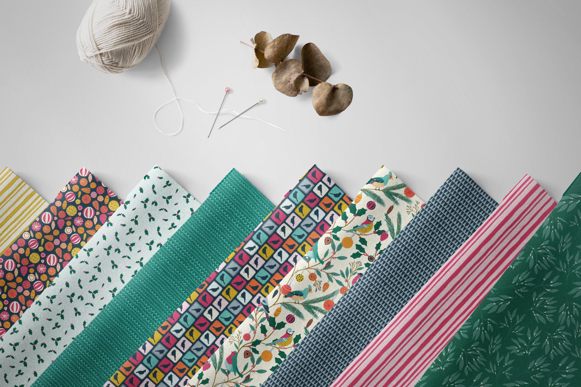 A fan arrangement of multiple patterned fabric or paper sheets in teal, pink, yellow, blue, and green tones on a light gray background, with a ball of yarn, sewing needles, and small dried leaves placed above them.
