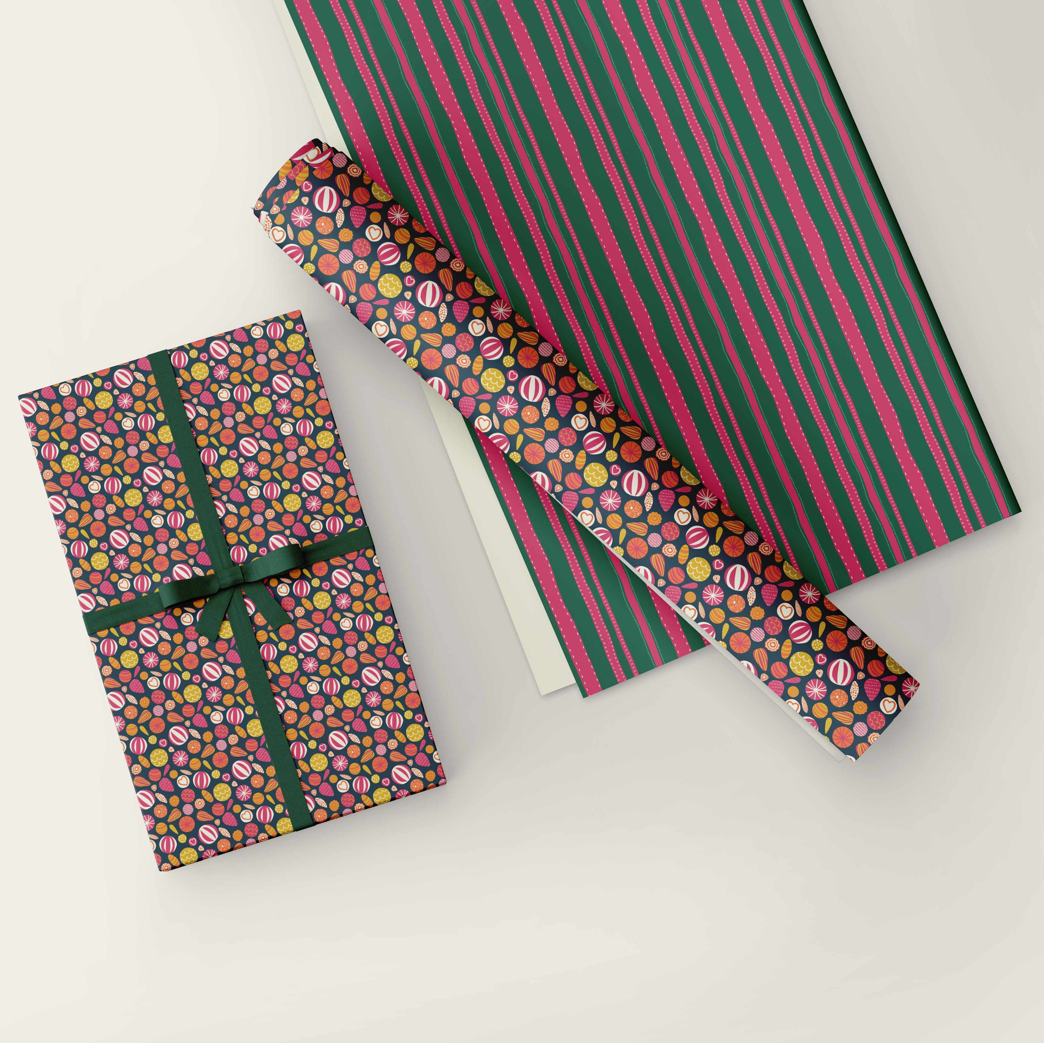 A gift-wrapping paper set shown on a light neutral background, including a wrapped rectangular box tied with a dark green ribbon, a rolled section of the patterned paper, and a larger flat sheet with vertical pink and dark green stripes.