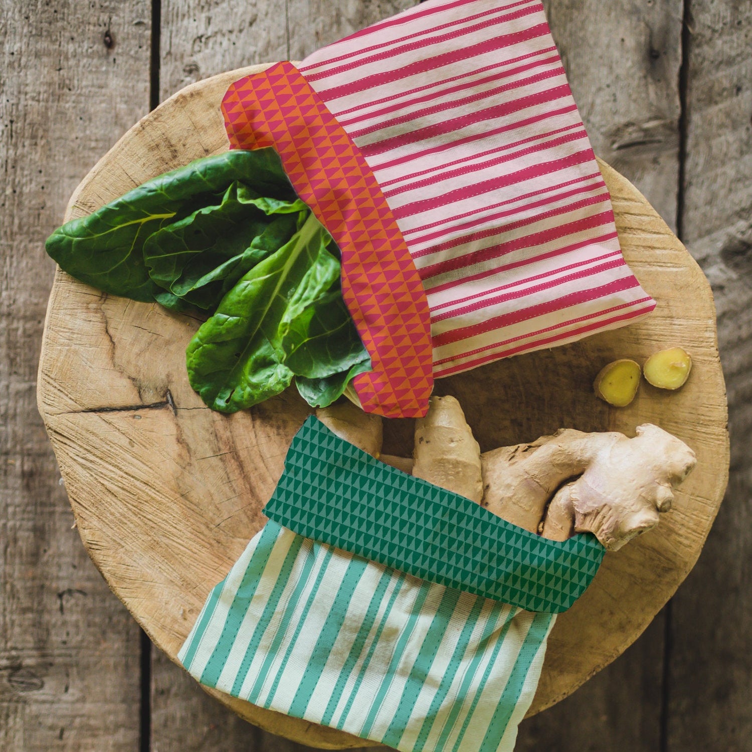 Two reusable fabric cloth pouches with striped patterns, one red and pink striped and one green and white striped, displayed on a round wooden cutting board with fresh greens, ginger, and garlic on a rustic wooden table.