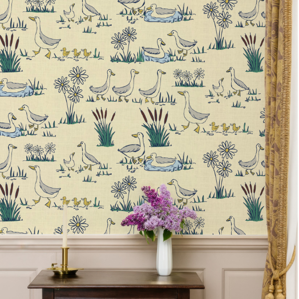 A wallpaper pattern featuring ducks, geese, and ducklings in muted blue, green, and yellow on a warm cream background, styled on a wall above a dark wood console table with a vase of purple lilacs.