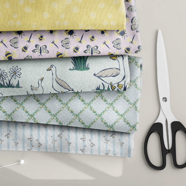 A stack of five patterned fabric bolts in soft pastel colors, arranged neatly on a light surface with a pair of black-handled scissors to the right.