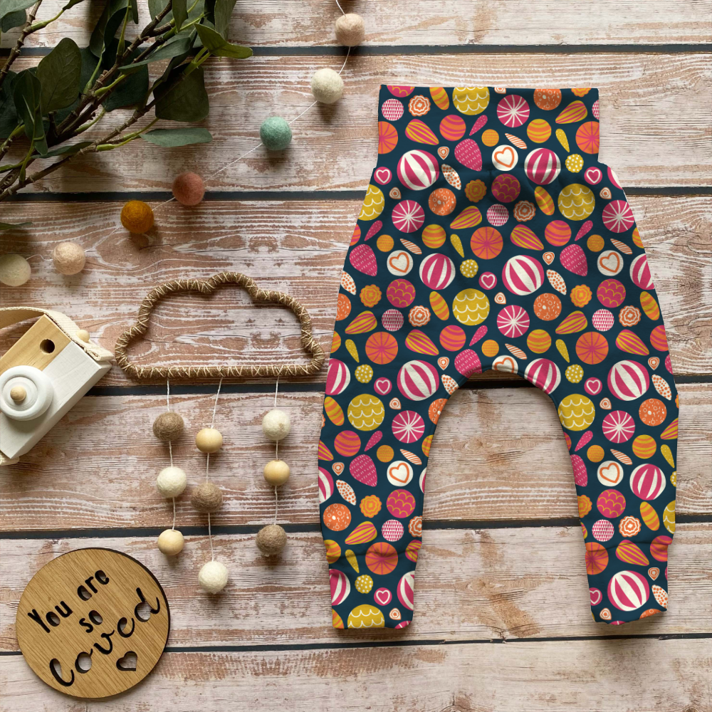 A pair of baby pants laid flat on a rustic wooden surface, covered in a colorful candy and dessert print on a dark navy background. The scene is styled with a wooden camera, a beaded garland, a wooden sign reading 'You are so loved', and greenery.