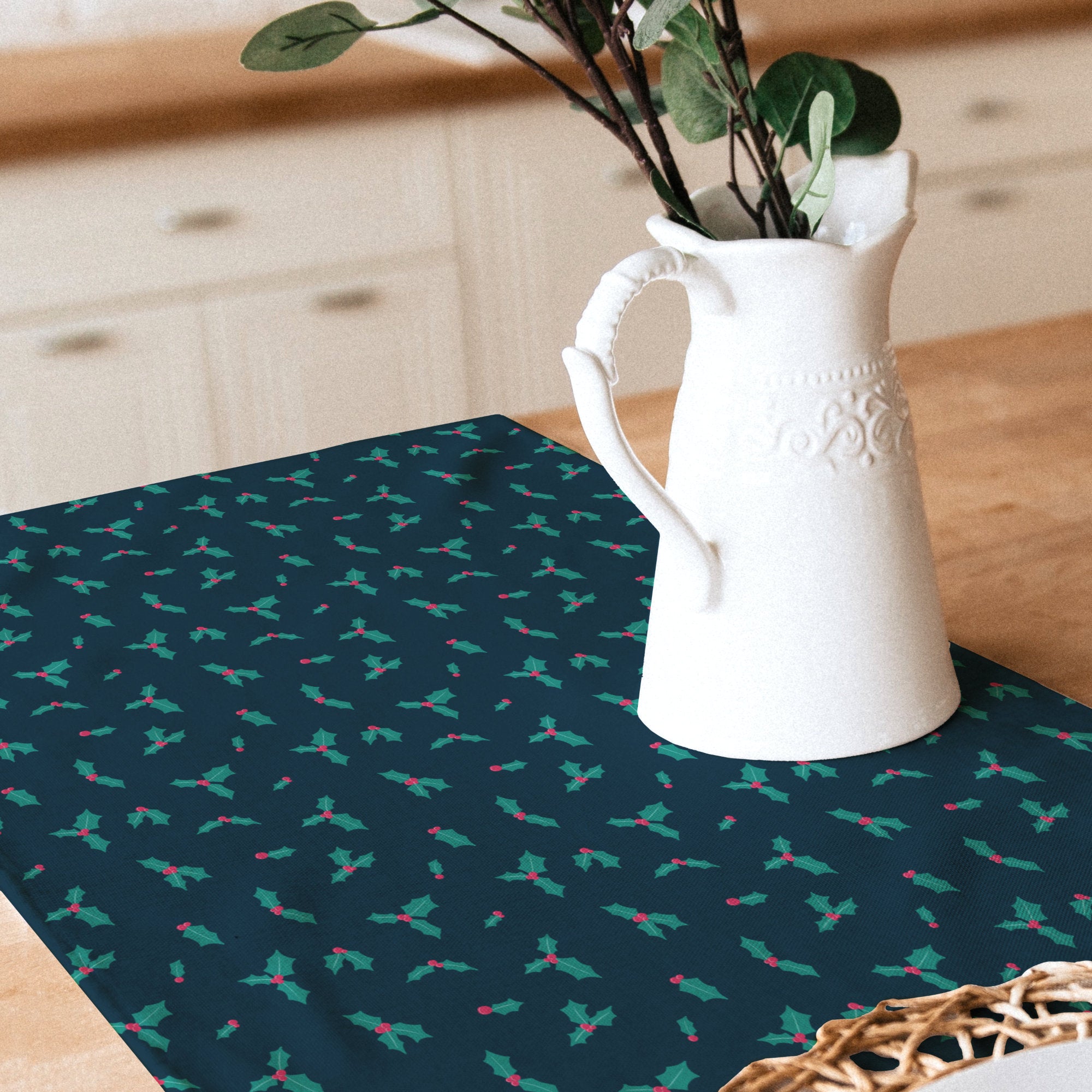 A dark blue table runner with a repeating holly and berry pattern, styled on a wooden table with a white ceramic pitcher and greenery in the background.