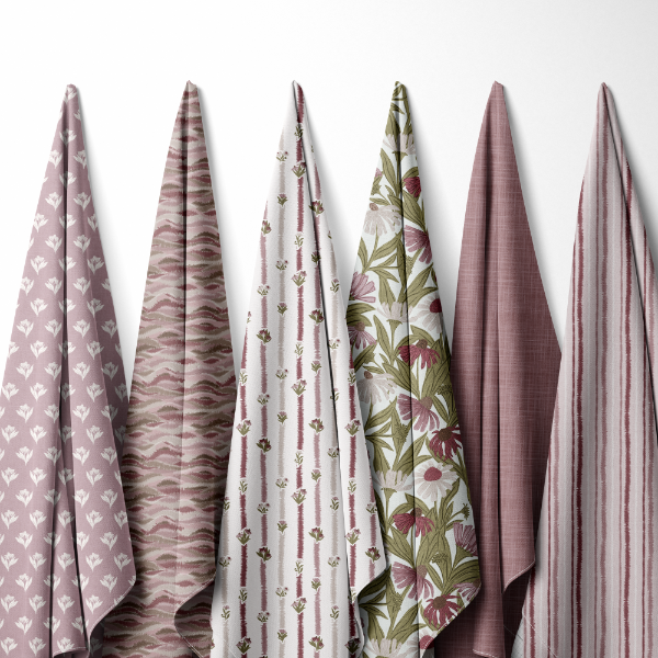 Six folded fabric cloths or napkins in muted pink, mauve, and floral tones hang vertically against a plain white wall. The fabrics show different prints including stripes, wavy lines, small floral motifs, and a larger botanical pattern.