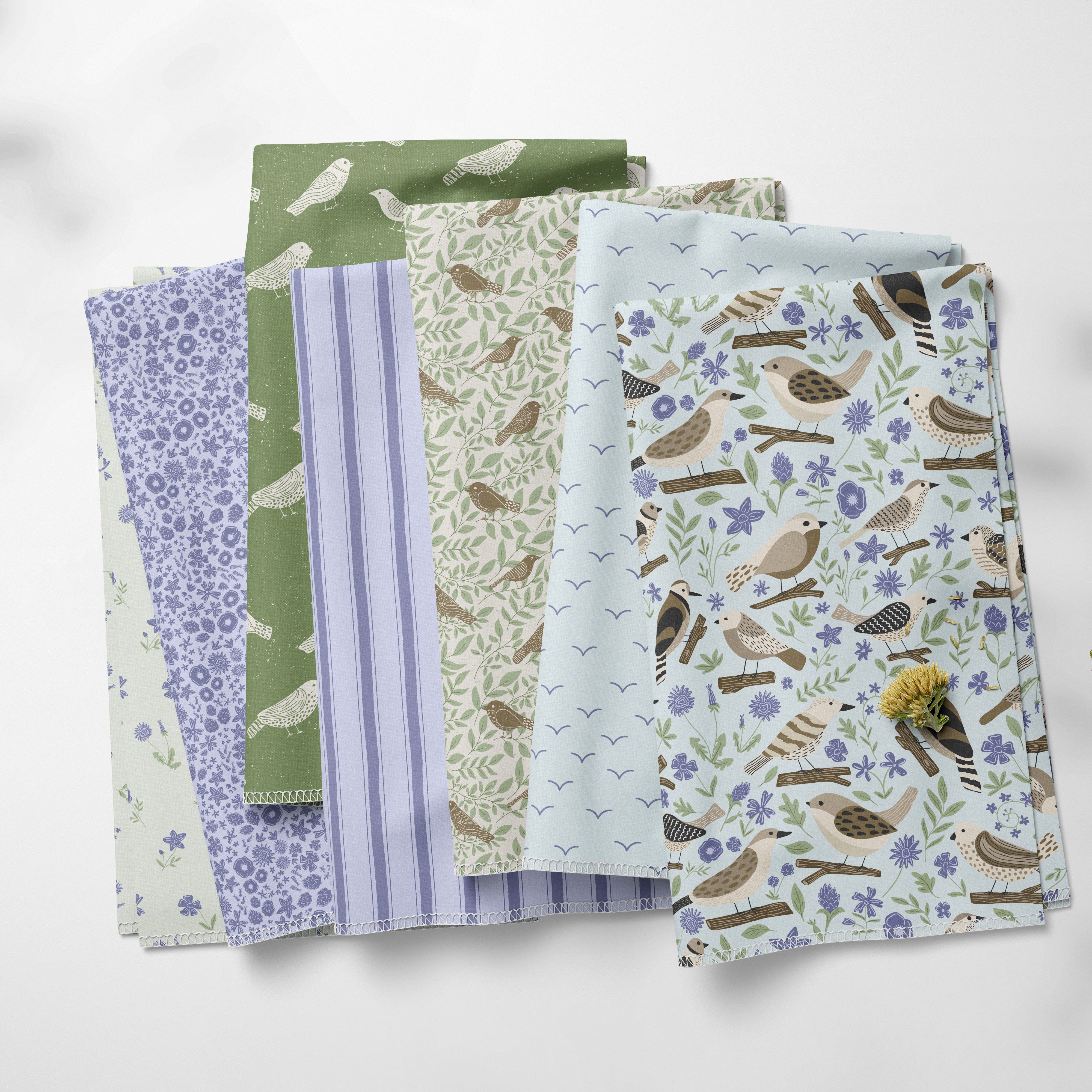 A stack of six patterned fabric swatches in soft pastel colors, including green, lavender, beige, and light blue, with bird and floral motifs, arranged on a light gray background with soft leaf shadows.