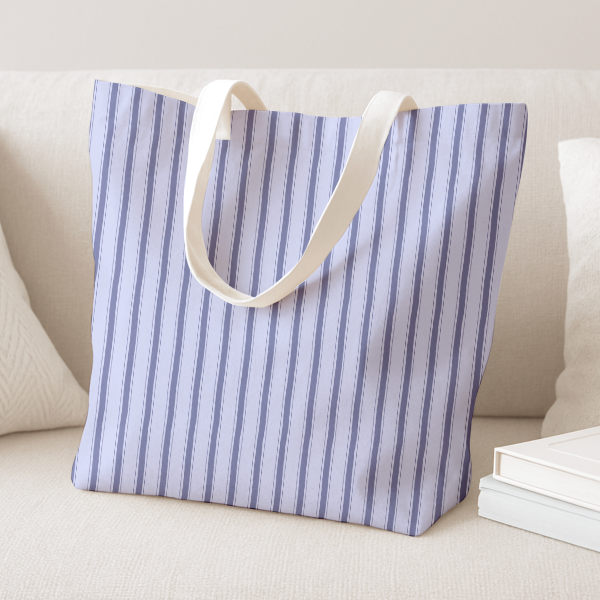 A tote bag with vertical blue and white stripes and cream-colored shoulder straps, displayed on a light beige sofa next to pillows and books.