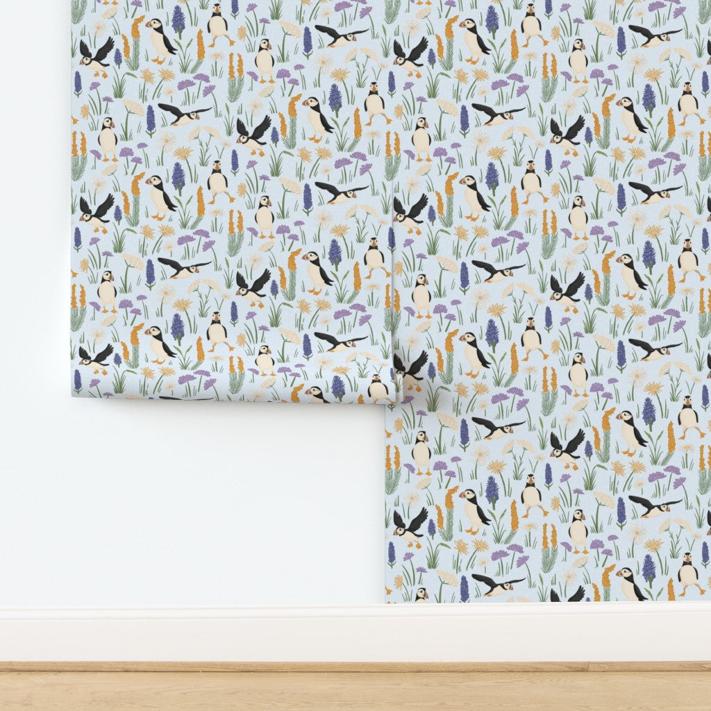 A roll of wallpaper with a repeating pattern of penguins, toucan birds, and purple flowers on a pale blue background, shown partially unrolled against a white wall and light wood floor.