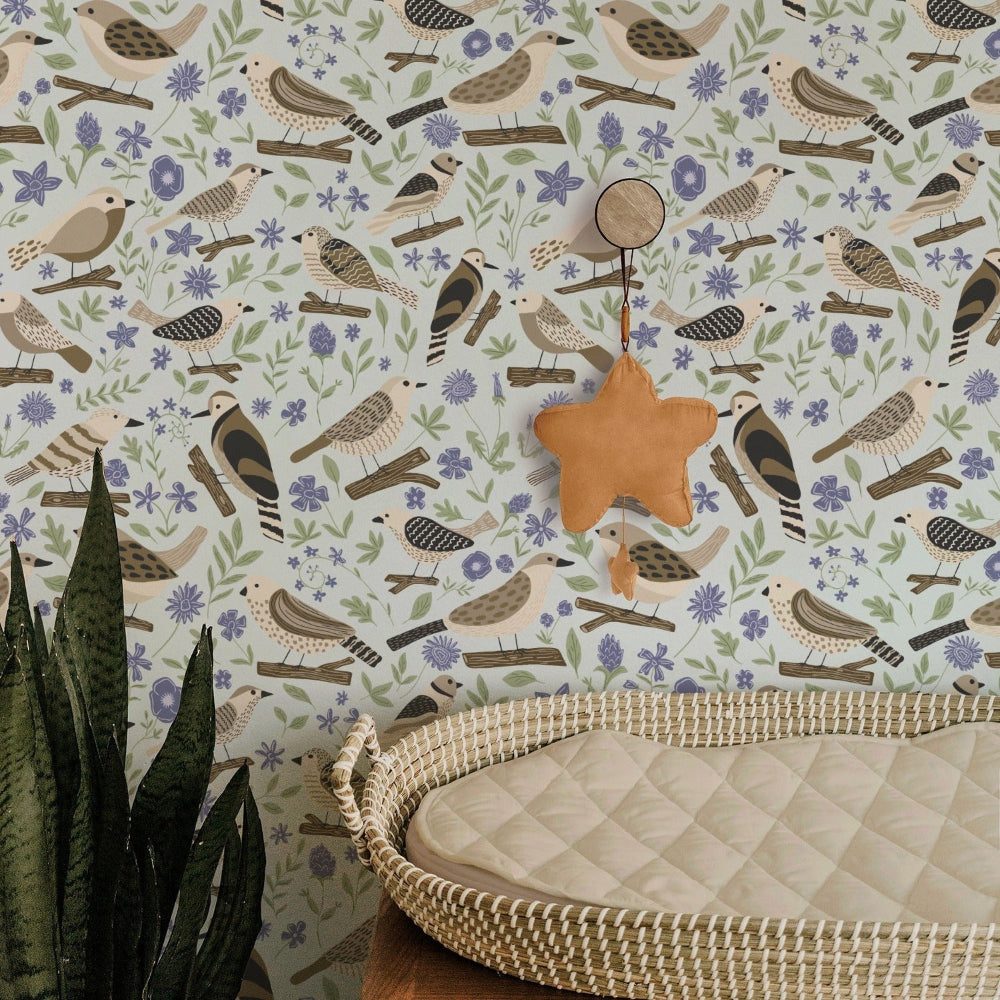 A styled nursery corner featuring wallpaper with a repeating pattern of birds, flowers, and leaves in muted blue, green, beige, and brown tones. A woven rattan bassinet with a quilted cream mattress is in the foreground, along with a hanging tan star ornament.