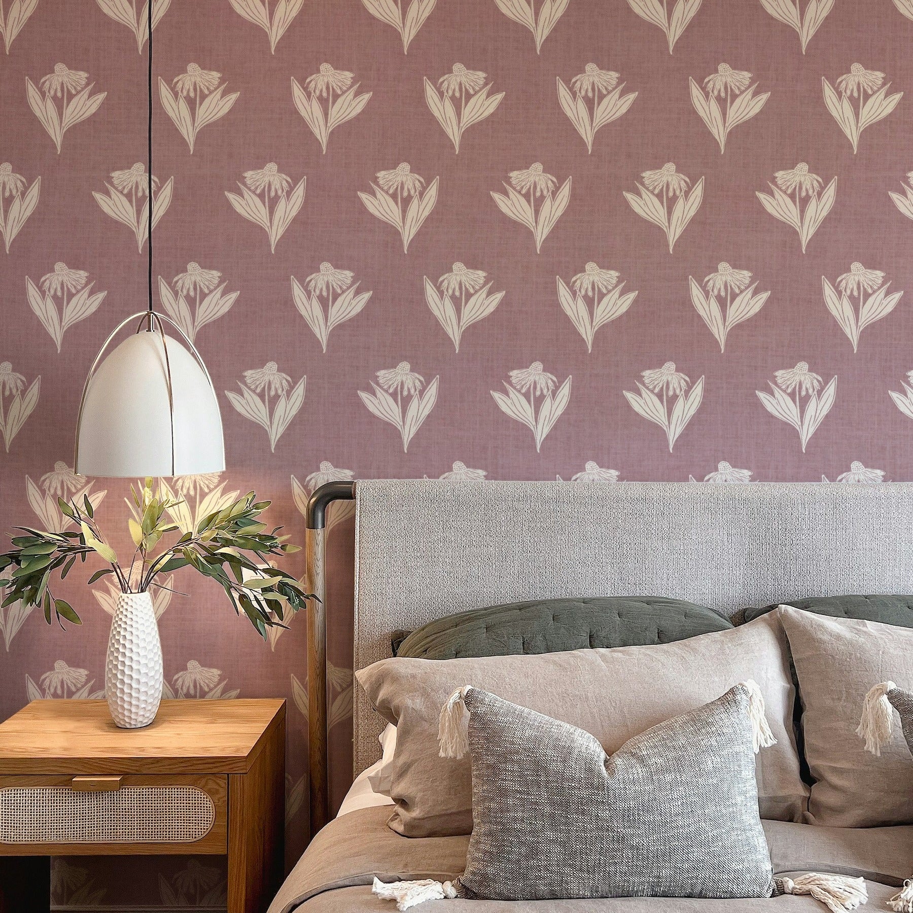 A styled bedroom corner featuring dusty mauve wallpaper with a repeating cream floral pattern, a wooden nightstand with a white pendant lamp and vase, and a bed with neutral-toned bedding and pillows.