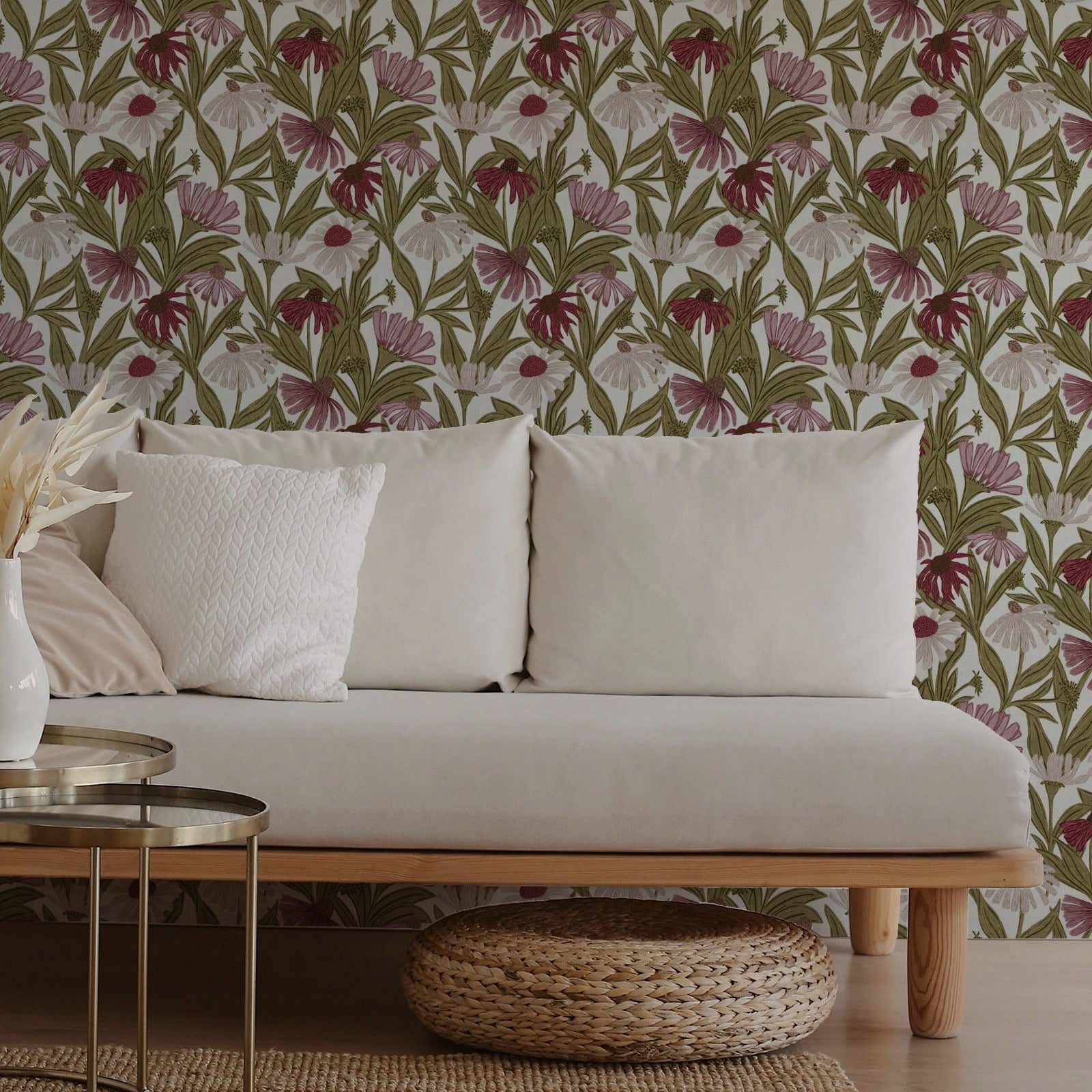 A styled interior scene showing a light beige sofa with white pillows against a wall covered in a botanical floral wallpaper with green leaves and pink flowers. A woven round ottoman sits in front of the sofa, with a small glass-top side table on the left.