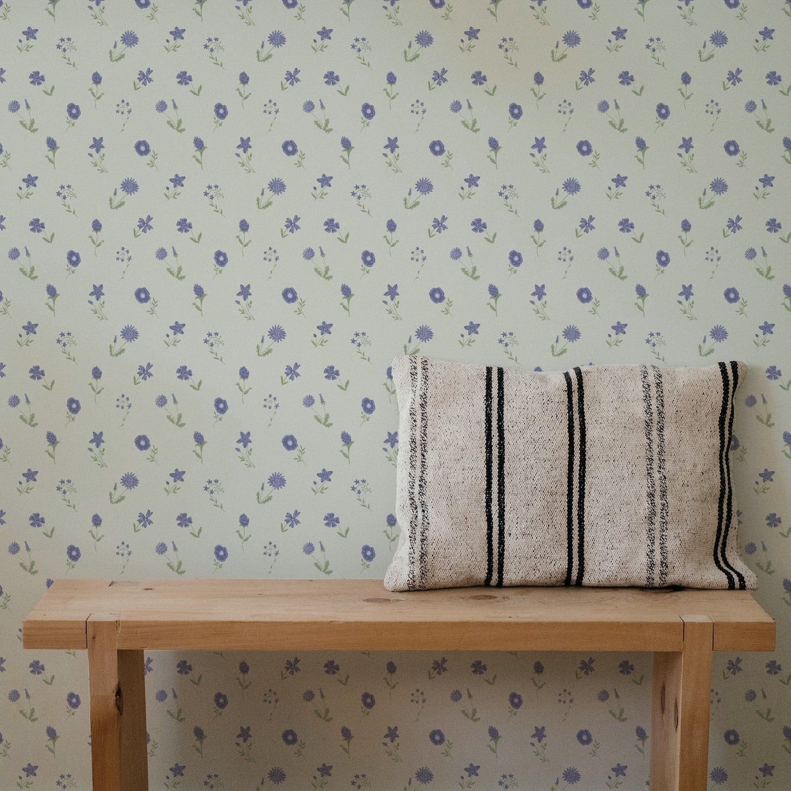 A light natural wood bench placed against a wall with repeating small floral wallpaper in muted blue and green. A rectangular woven throw pillow with black vertical stripes rests on the bench.