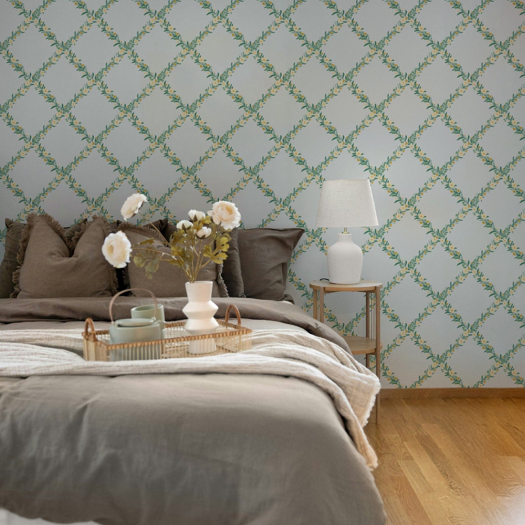 A styled bedroom interior featuring a bed with taupe bedding, a small side table with a white lamp, and a wall covered in light blue wallpaper with a green trellis pattern.