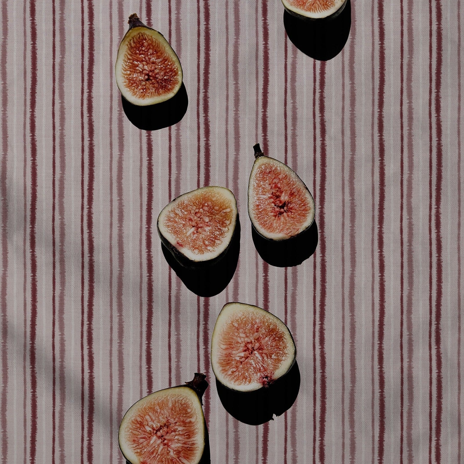 Six halved fresh figs with pinkish-red interiors and pale green skins are arranged on a vertically striped fabric background with dappled shadows.