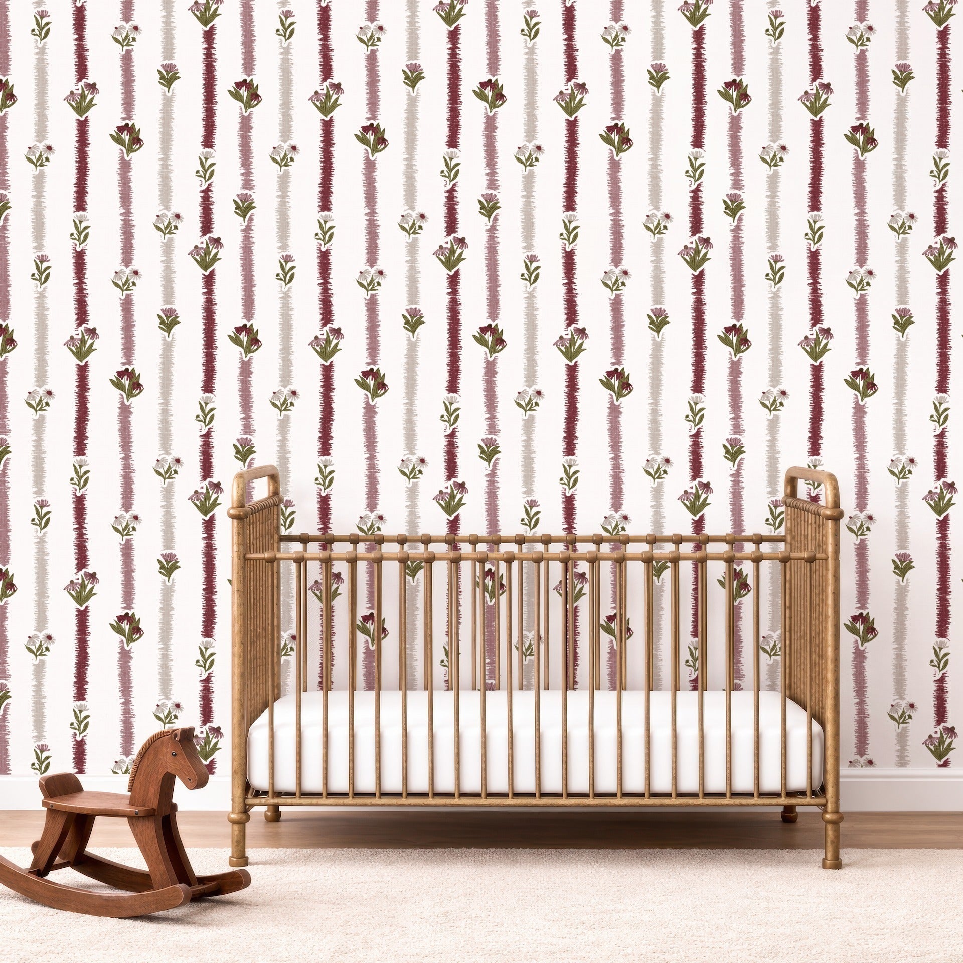 A nursery room scene featuring a gold metal crib with a white mattress, set against a wall with vertical burgundy and olive green floral patterns on a white background. A wooden rocking horse is on the floor to the left.