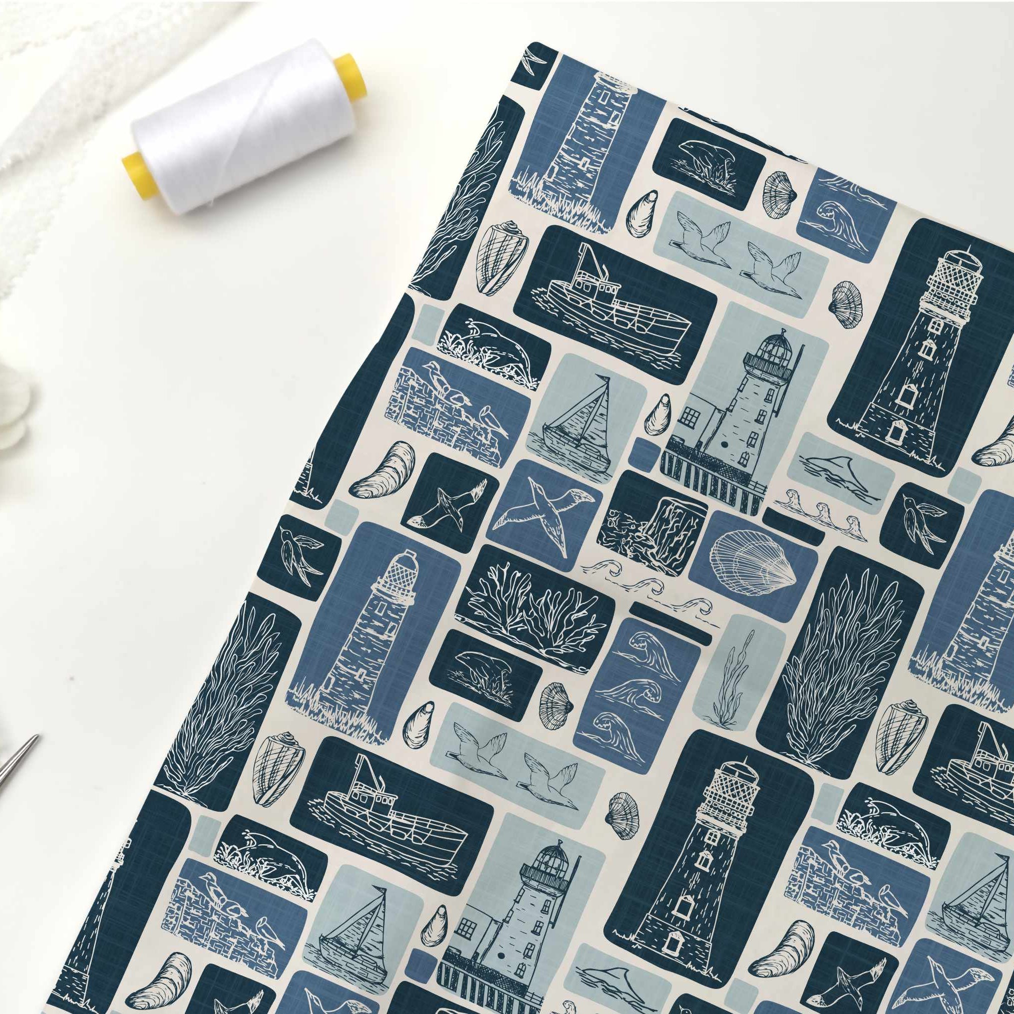 A flat-lay view of a fabric yardage featuring a nautical-themed print with lighthouses, sailboats, seashells, and starfish in shades of blue and navy on a white background. The fabric is styled on a white surface alongside a spool of white thread, silver scissors, and white flowers.