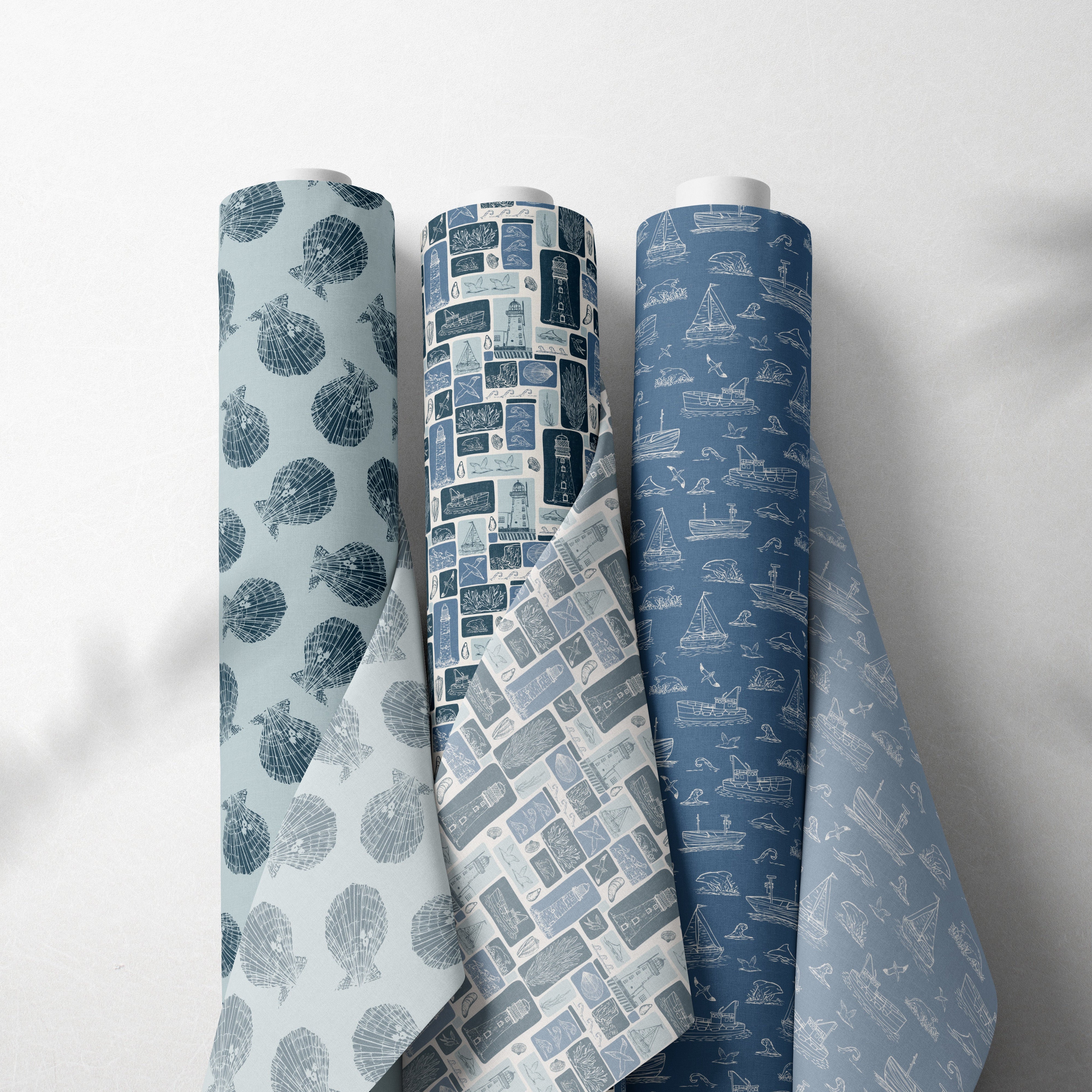 Three rolls of patterned wallpaper standing upright against a light gray wall with soft leaf shadows. The left roll has a light gray background with dark blue tree silhouettes, the middle roll shows a blue and gray geometric pattern, and the right roll has a medium blue background with white architectural line drawings.