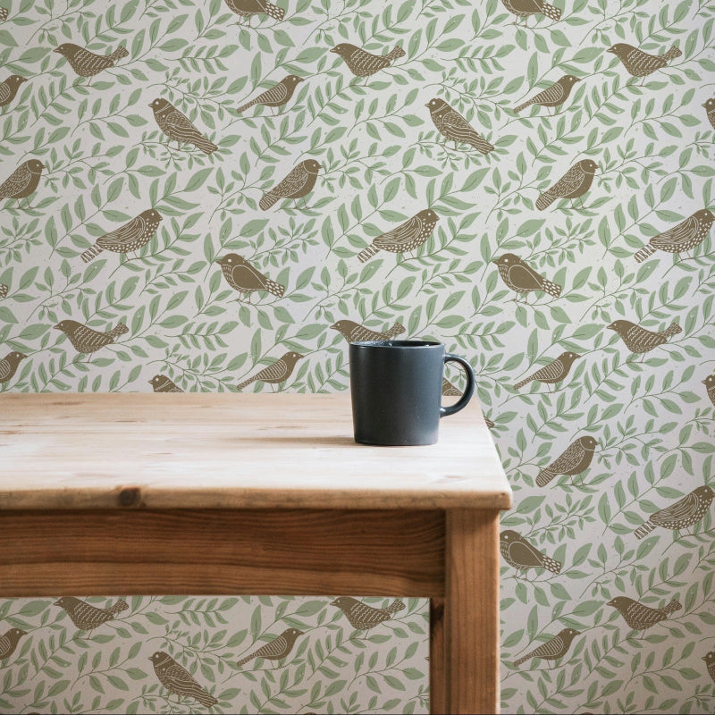 Green and brown bird and leaf patterned wallpaper shown on a wall behind a light wood table with a dark gray mug on top.