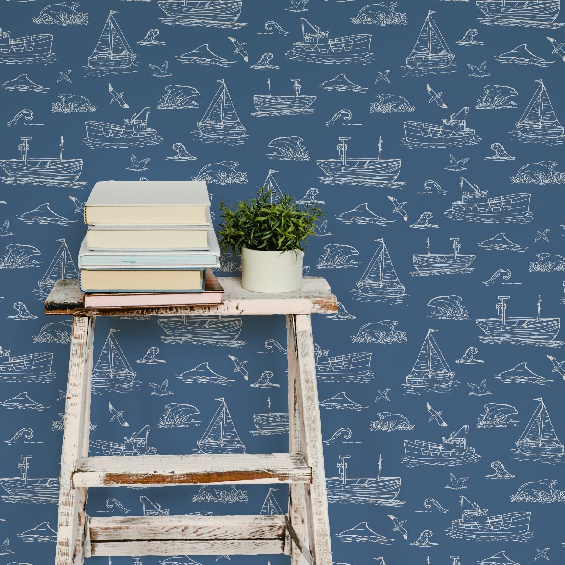 Nautical-themed wallpaper featuring a repeating pattern of white hand-drawn sailboats, ships, and sea life on a slate blue background, styled with a rustic white wooden stool holding books and a small potted plant.