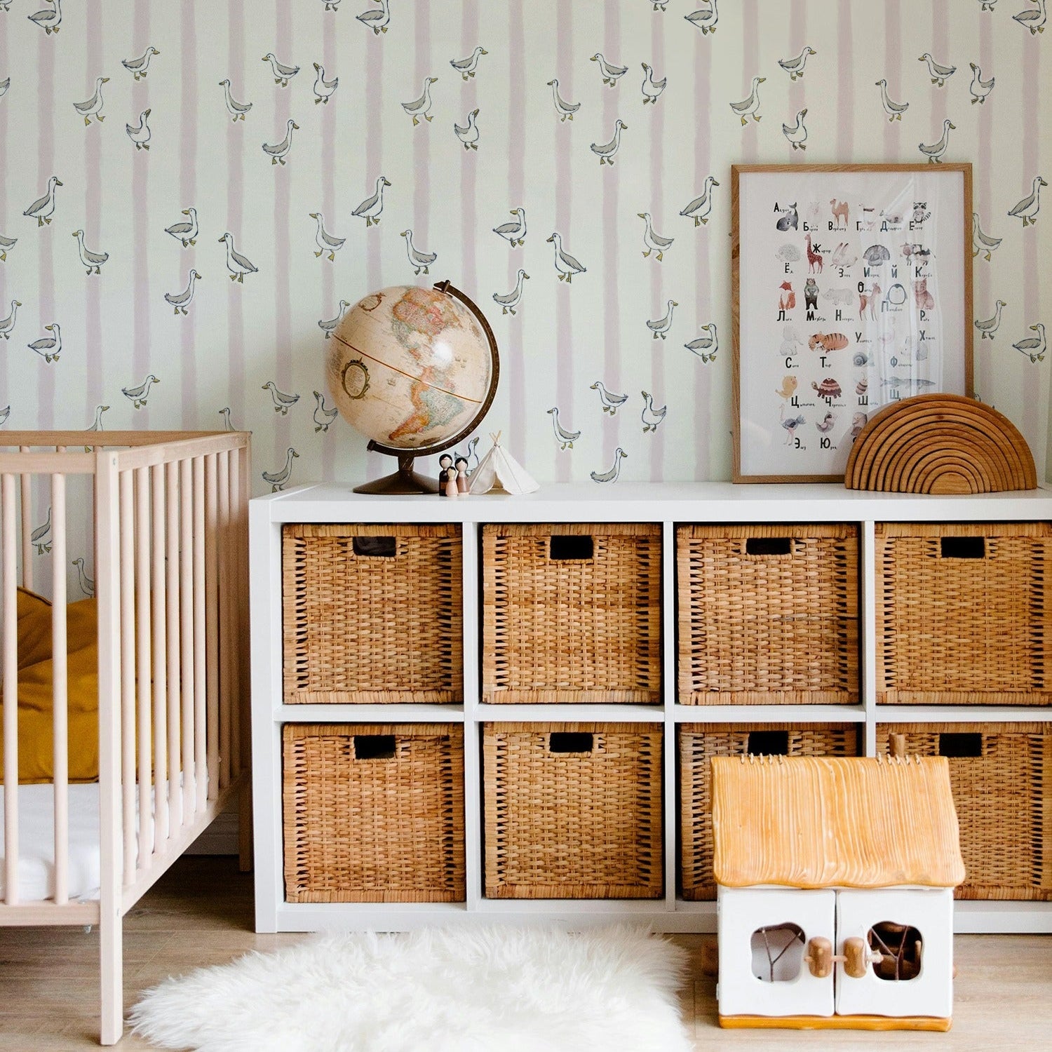 A styled nursery corner featuring a white storage cabinet with nine woven wicker baskets, a striped wallpaper with bird motifs, a wooden crib, a globe, and a small wooden house on a white faux fur rug.