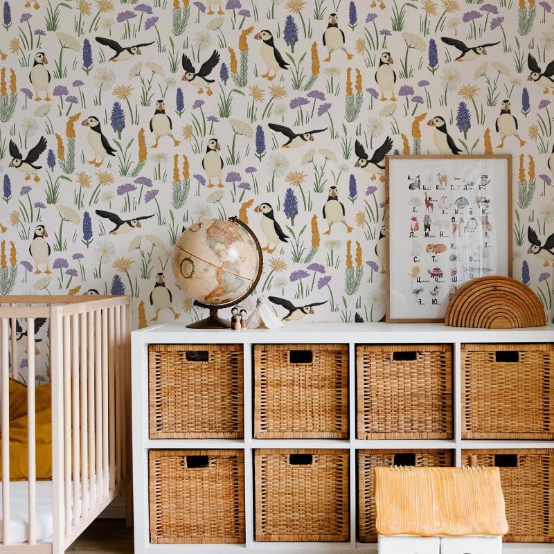 A styled nursery corner featuring a white storage cabinet with woven wicker bins, a patterned wallpaper with birds and reeds, a crib, a globe, and a small wooden house on a light wood floor.