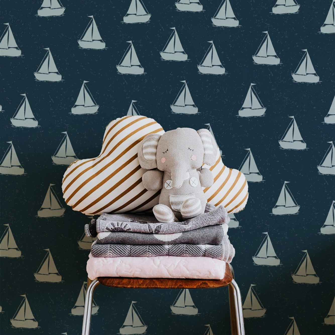 A styled nursery scene featuring a stack of folded baby textiles on a small stool in front of a dark navy wallpaper patterned with white sailboats. The stack includes striped heart-shaped pillows, a gray blanket, a pale pink towel, and a small plush elephant toy.