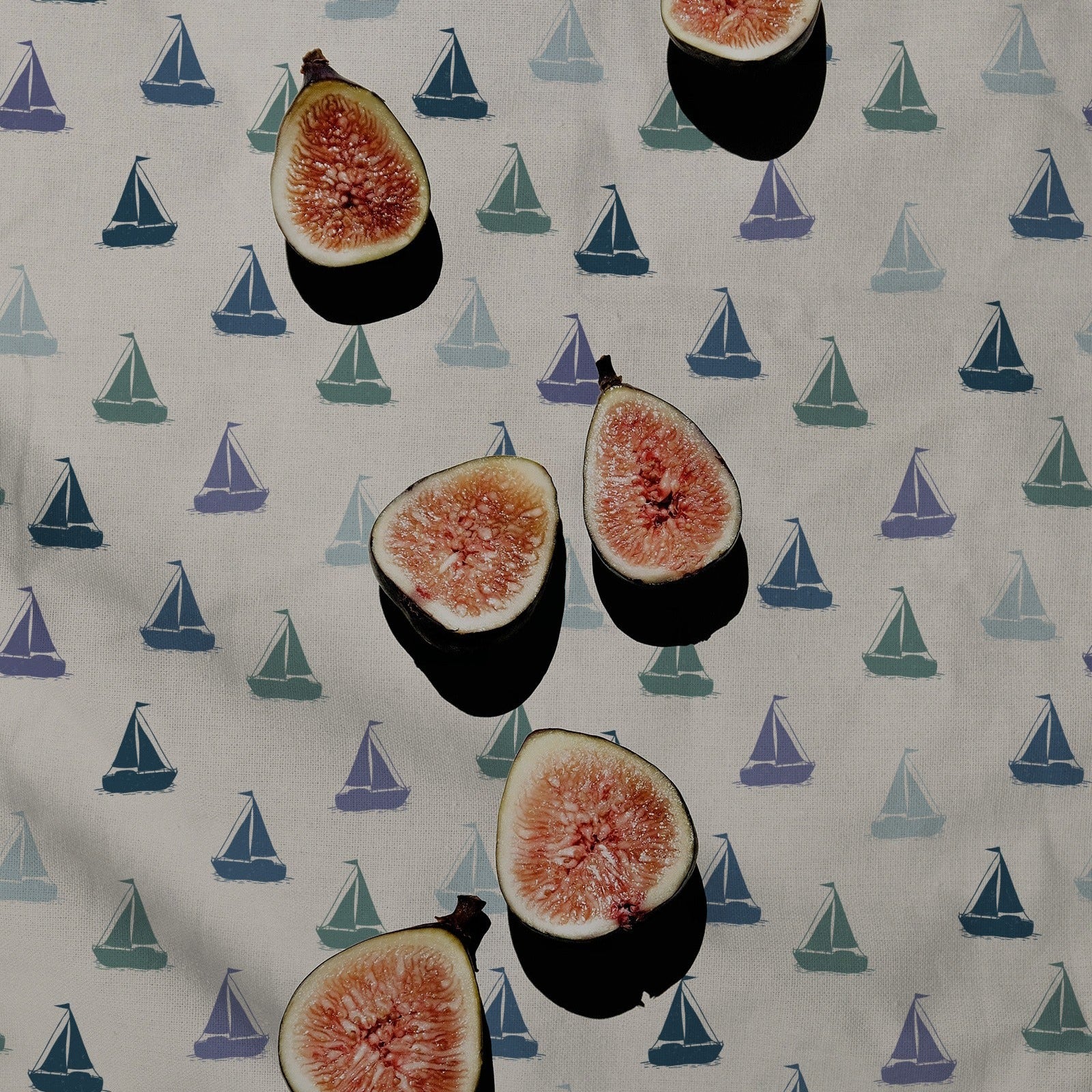 A close-up of a fabric surface with a repeating pattern of small sailboats in blue, teal, and purple on an off-white background, overlaid with several cut fig halves with dark shadows.