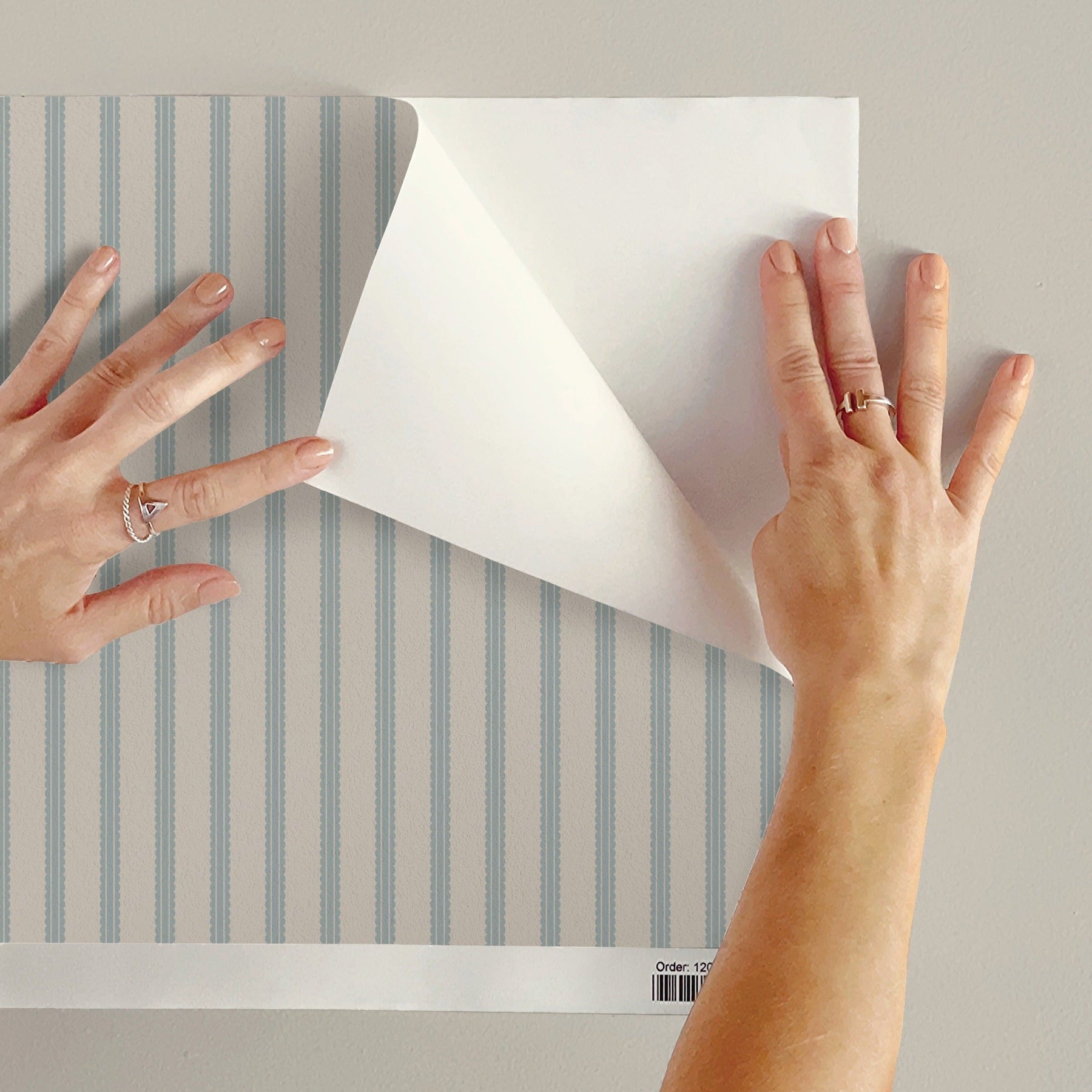 A person's hands peel back a corner of a sheet of wallpaper with vertical light blue and white stripes, showing the white backing paper. The background is a solid light gray wall.